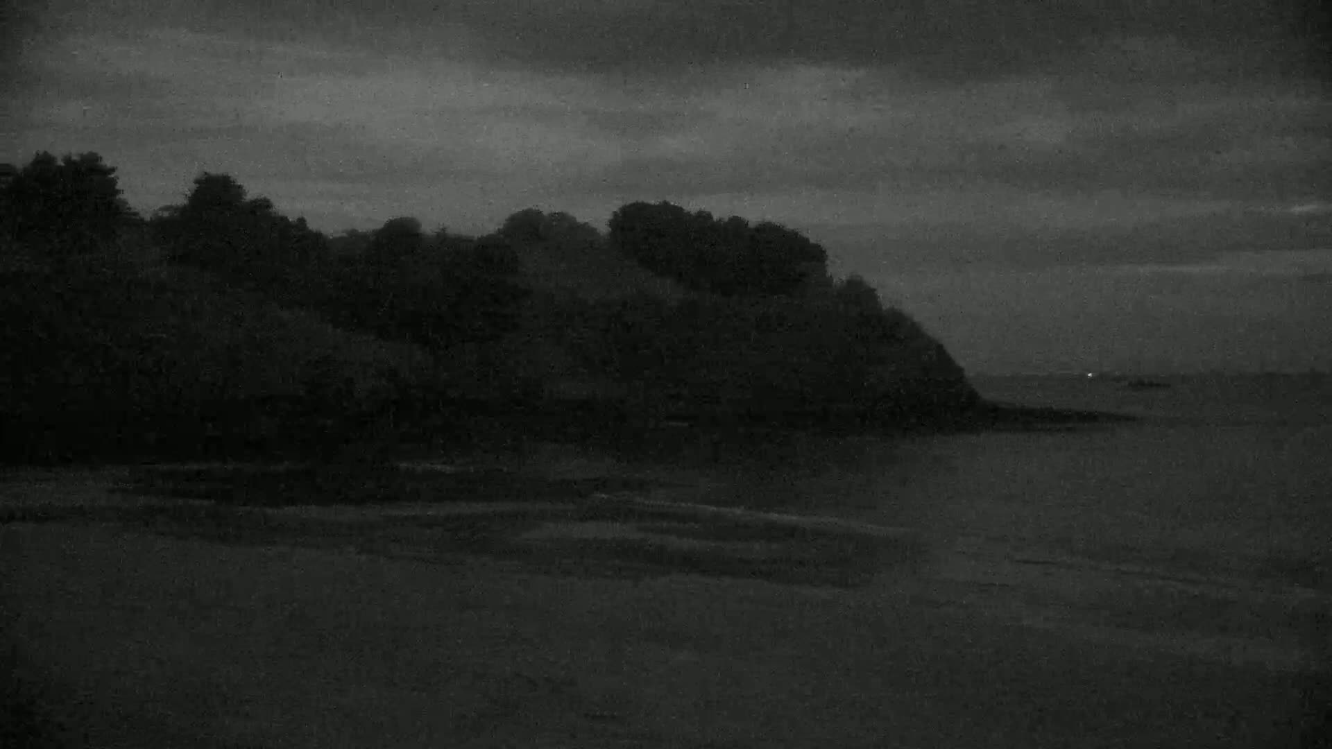 A dark, overcast sky looms over a beach and a silhouetted coastline with trees.