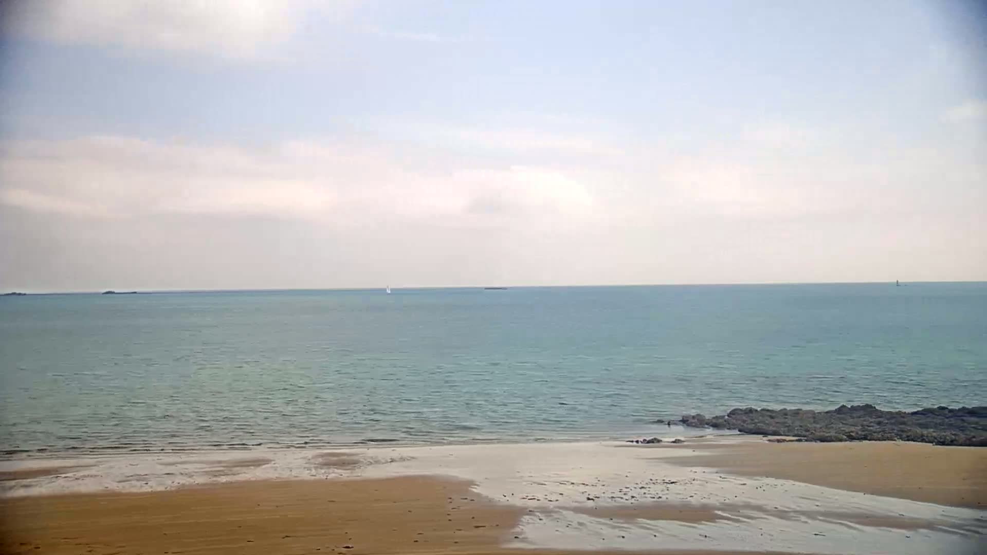 A sandy beach slopes gently down to a calm, light teal sea under a mostly cloudy sky.