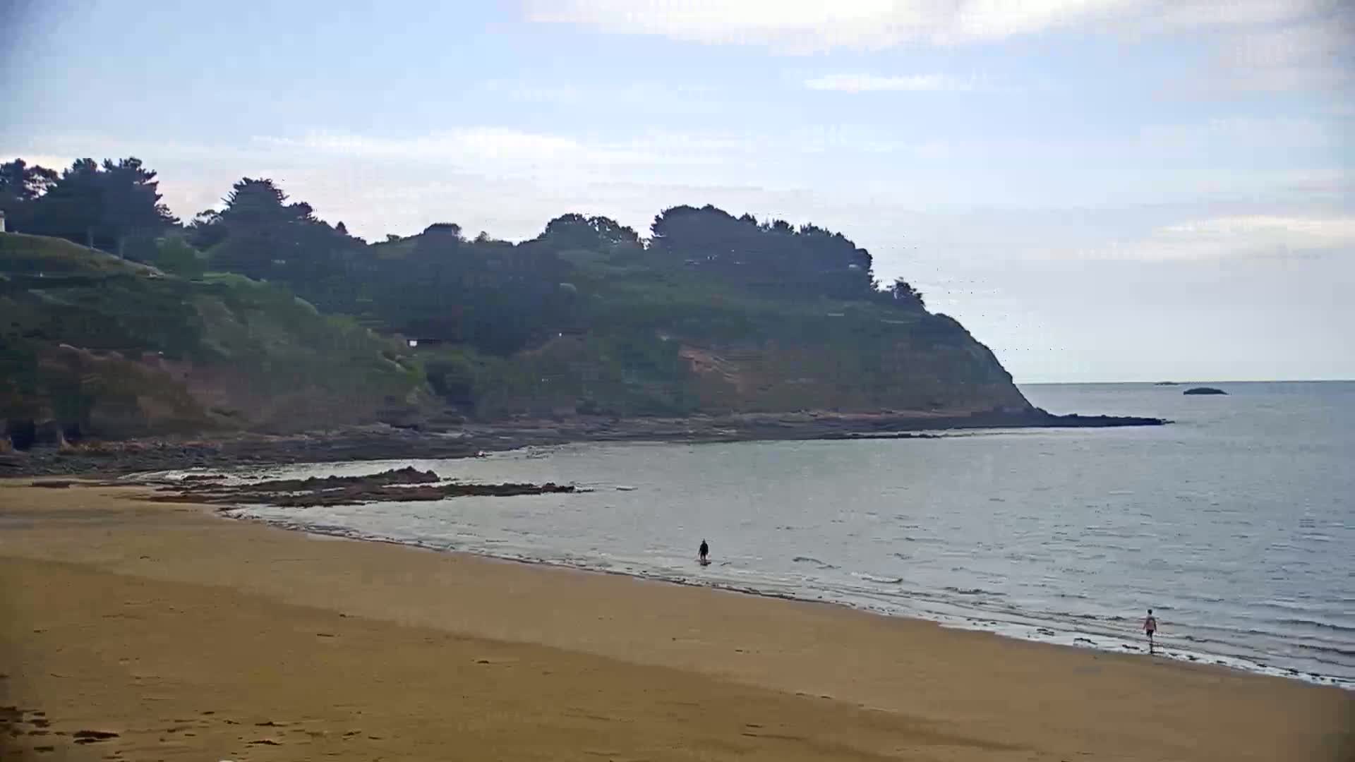 A sandy beach stretches out to calm, grey water lapping a rocky shoreline beneath a mostly clear, light-grey sky, with dark green vegetation covering the cliffs behind.