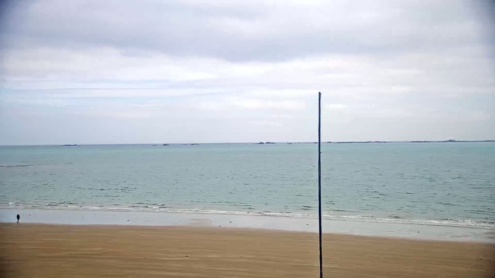 A lone person walks on a sandy beach beside a calm, light-green sea under an overcast sky.