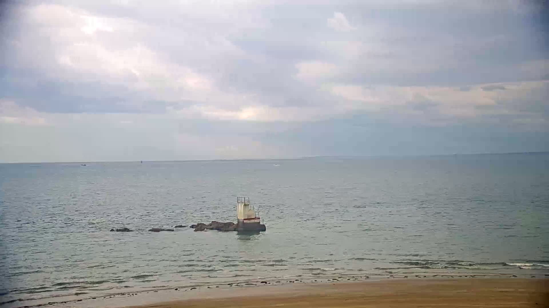 A small, white structure sits on rocks in a calm, grey sea under a cloudy sky, with a sandy beach in the foreground.