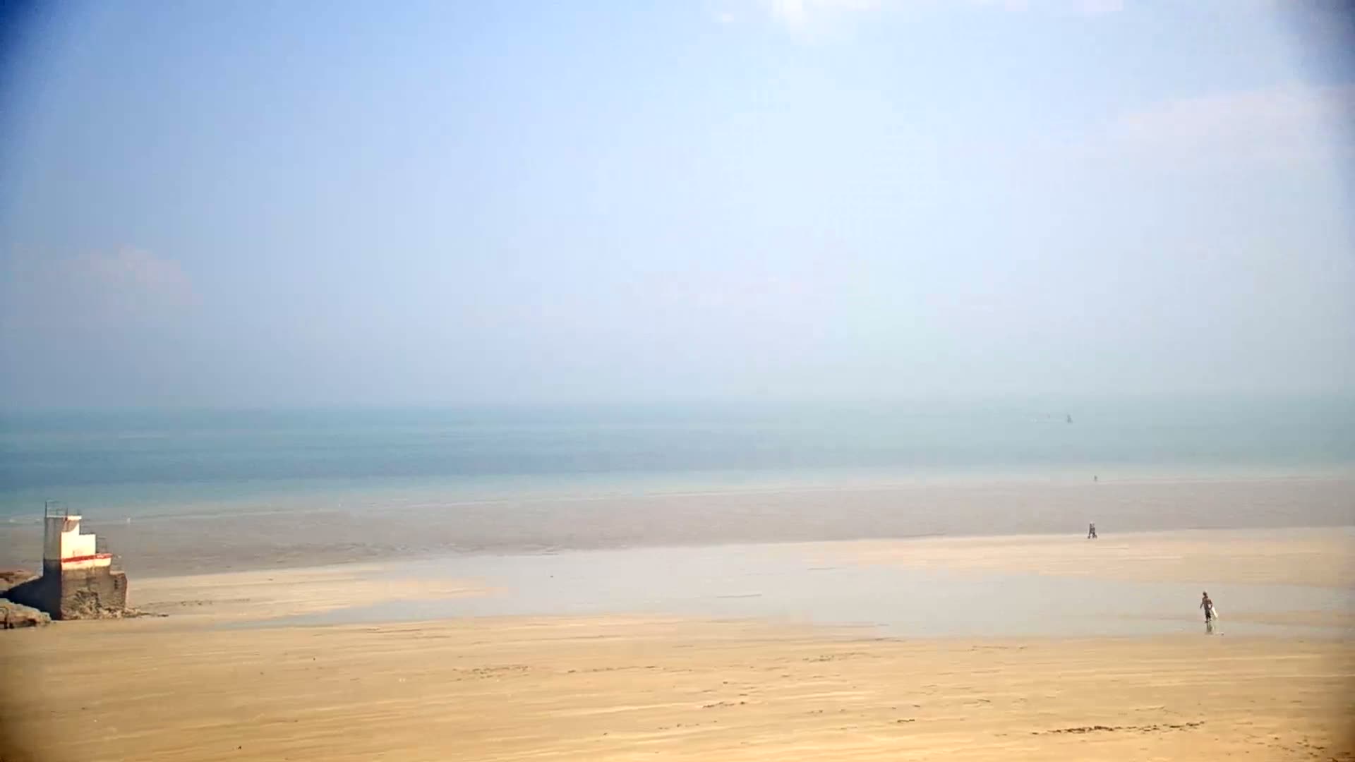 A hazy, light-blue ocean meets a broad, sandy beach where a few people walk near a small, white structure on the shore.