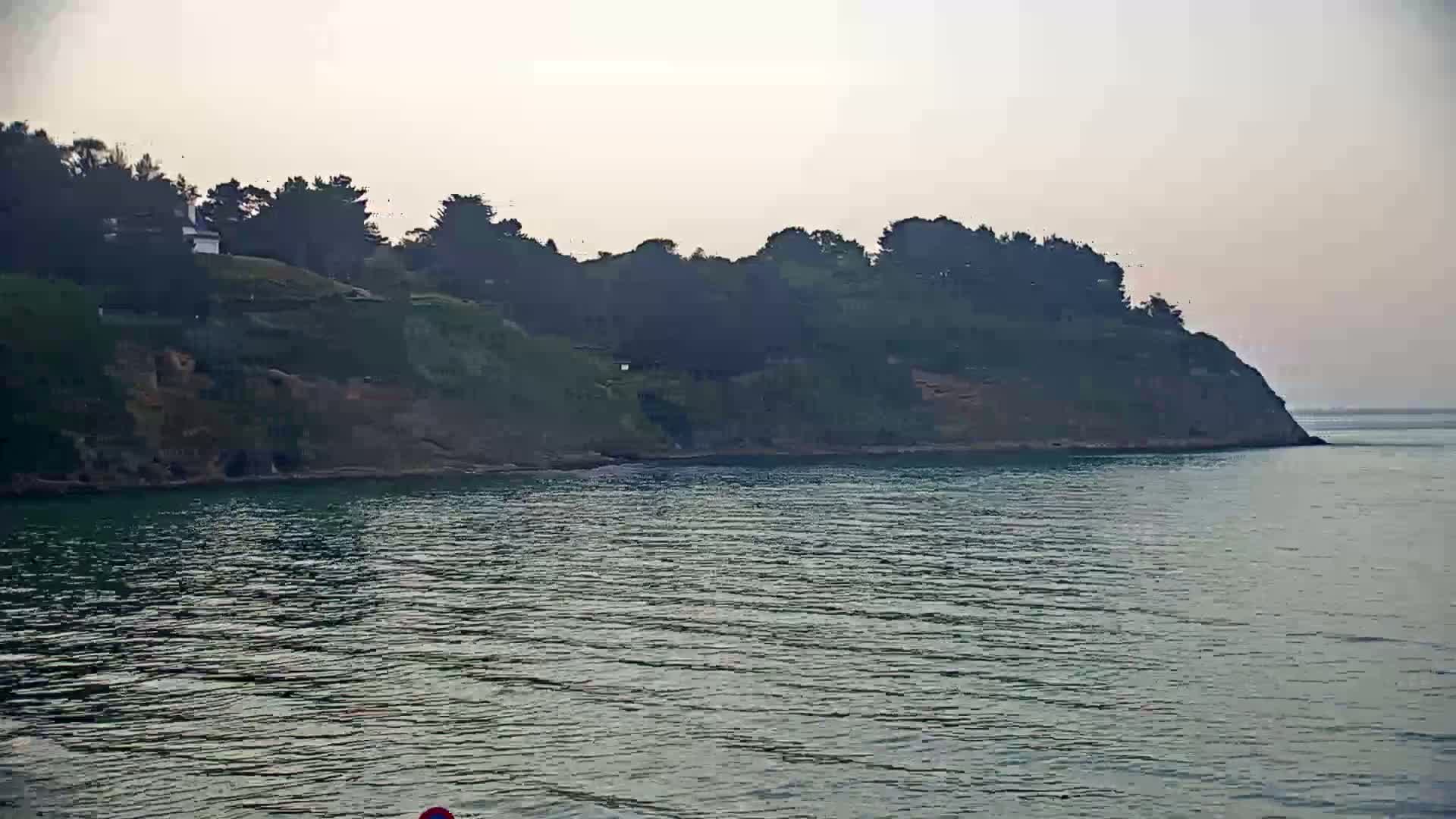 A verdant, tree-covered cliff overlooking calm, dark-green water under a hazy sky.