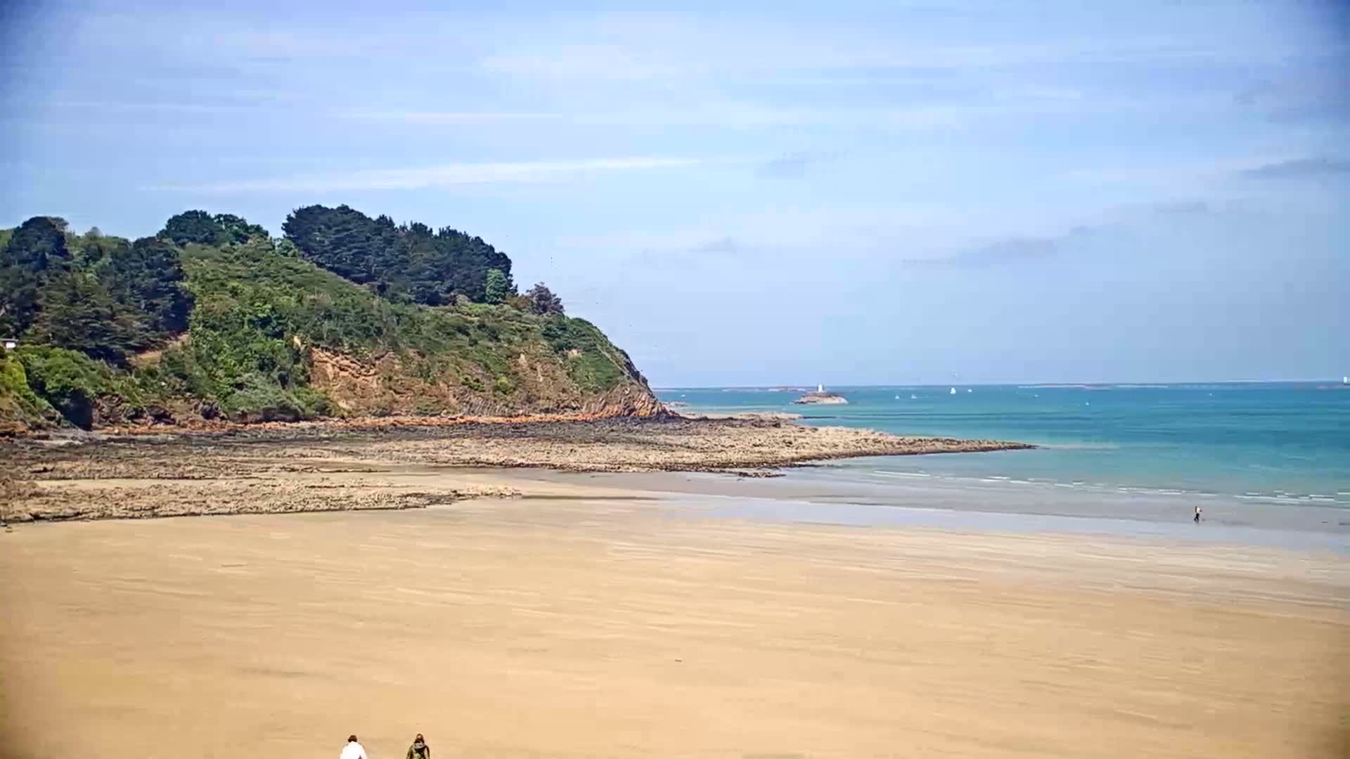 A sandy beach stretches to a calm, teal ocean under a clear blue sky, with a rocky outcrop and green hillside in the background.