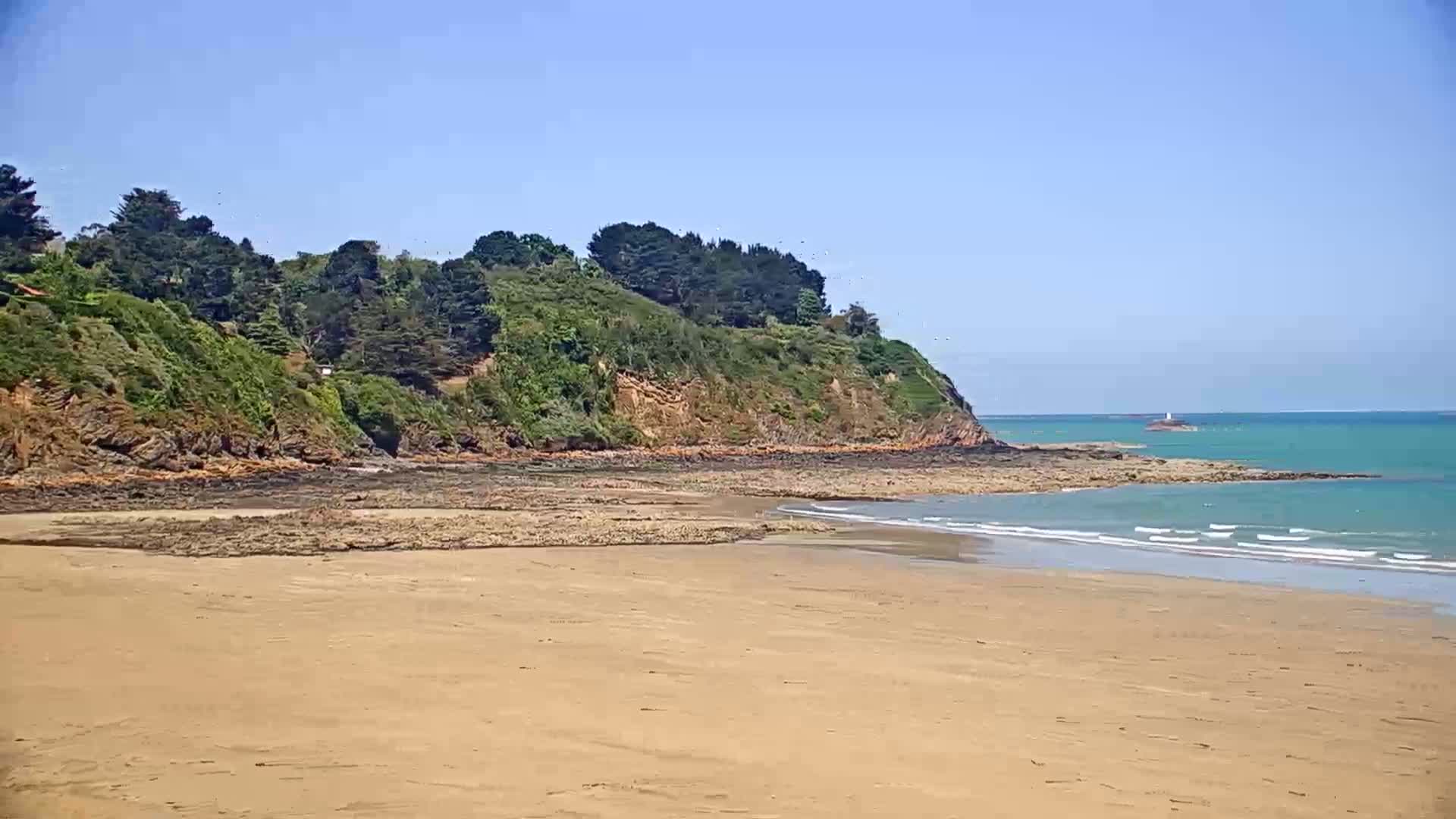 A sandy beach stretches out to a calm sea, meeting a rocky shoreline topped with lush green vegetation under a clear blue sky.