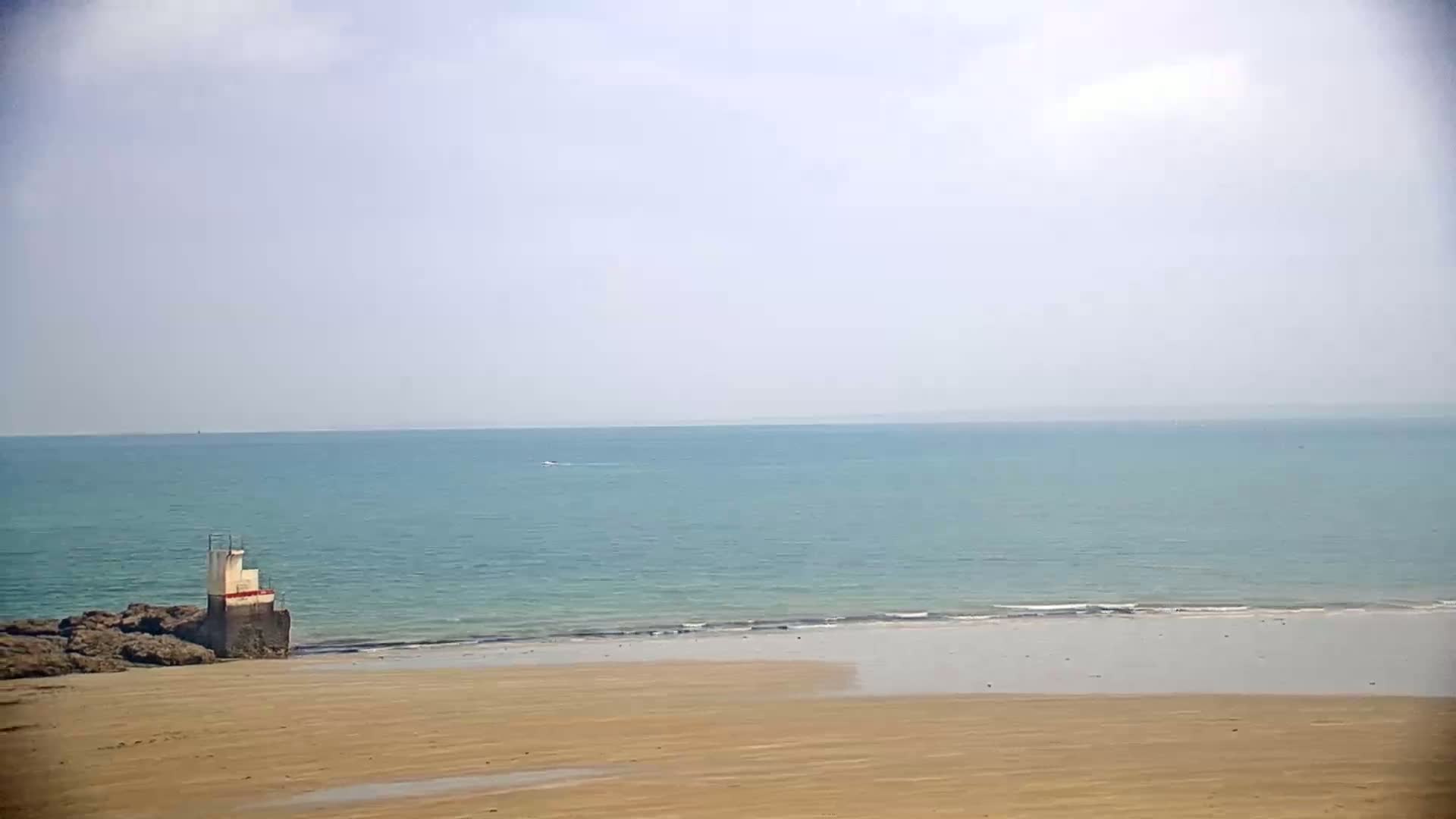 A small, white structure sits on a rocky outcrop near a sandy beach and a calm, light-blue ocean under a mostly cloudy sky.