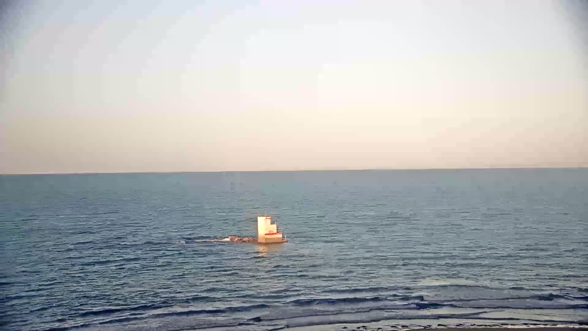 A small, light-colored structure sits on the calm, blue ocean under a pale sky.