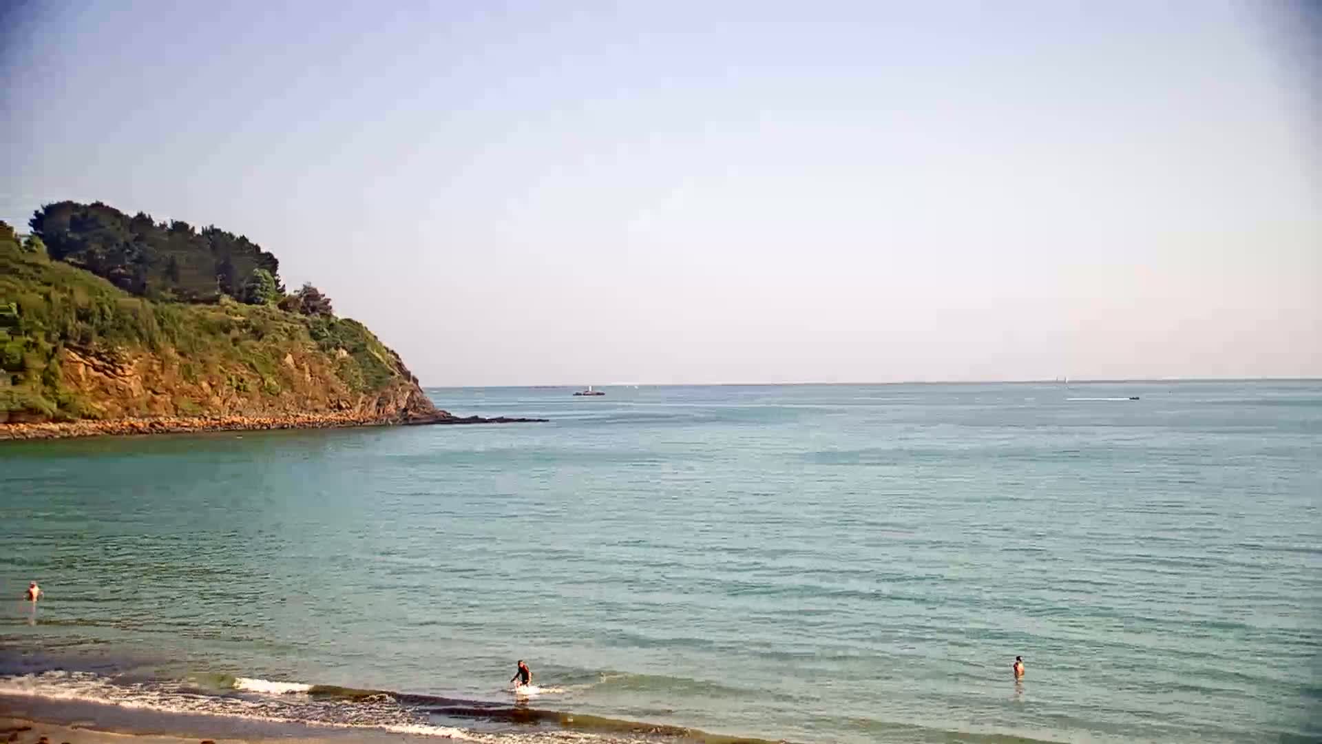A calm, clear day shows a sandy beach with a few people swimming in the light teal water near a small, rocky, green-covered headland.