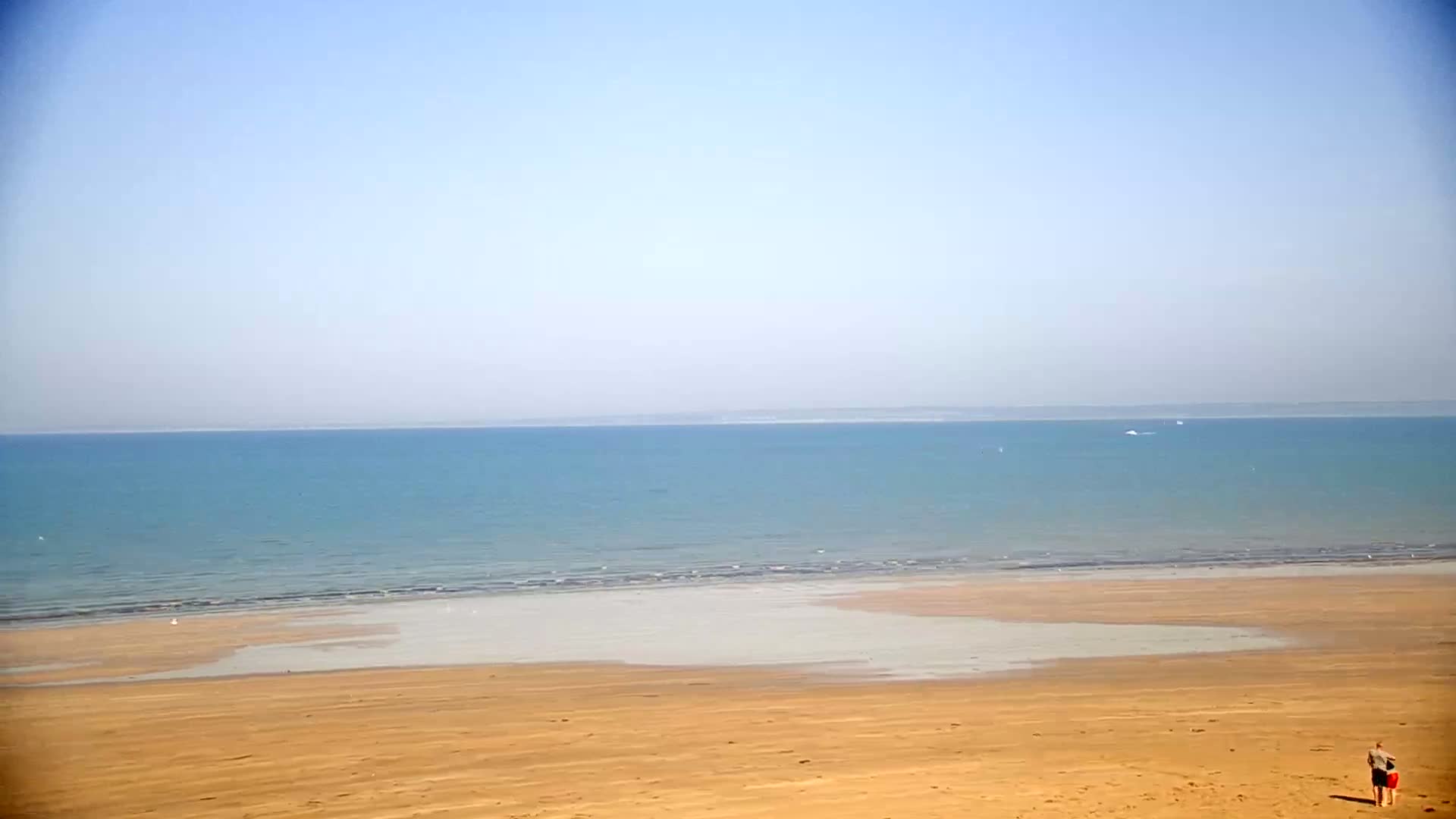 A calm, light-blue ocean meets a wide, sandy beach under a clear, pale-blue sky.