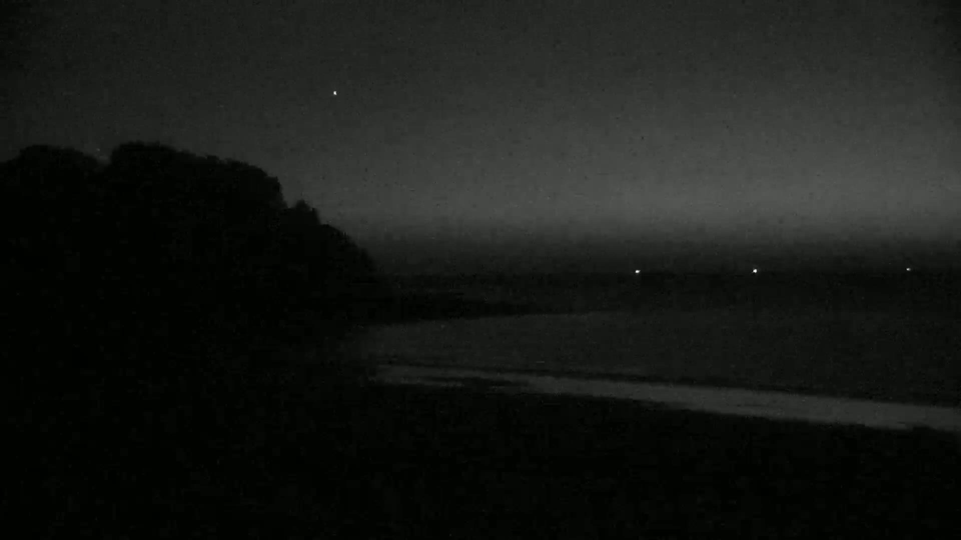 A dark, nighttime scene shows a silhouetted shoreline with a calm body of water under a slightly hazy sky.