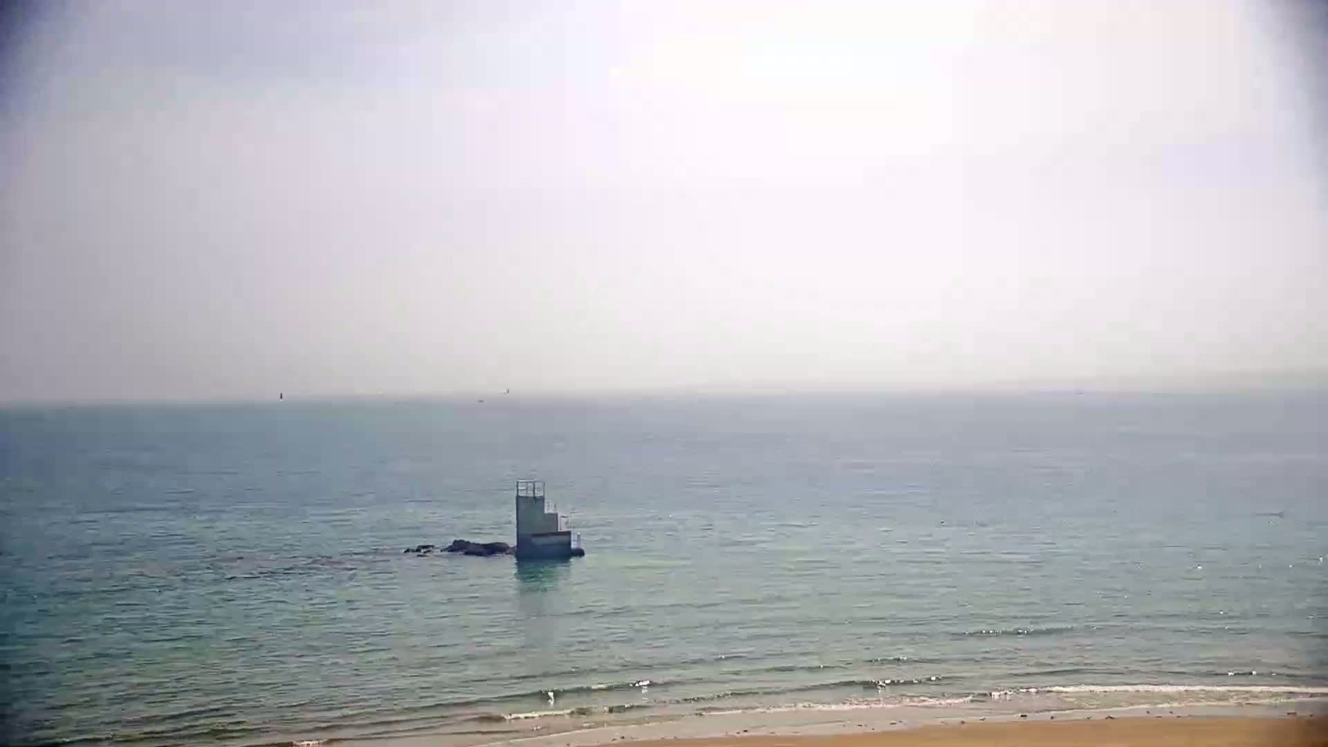 A hazy, light-colored beach with calm ocean waves shows a small, square diving platform on a rocky outcropping in the water.