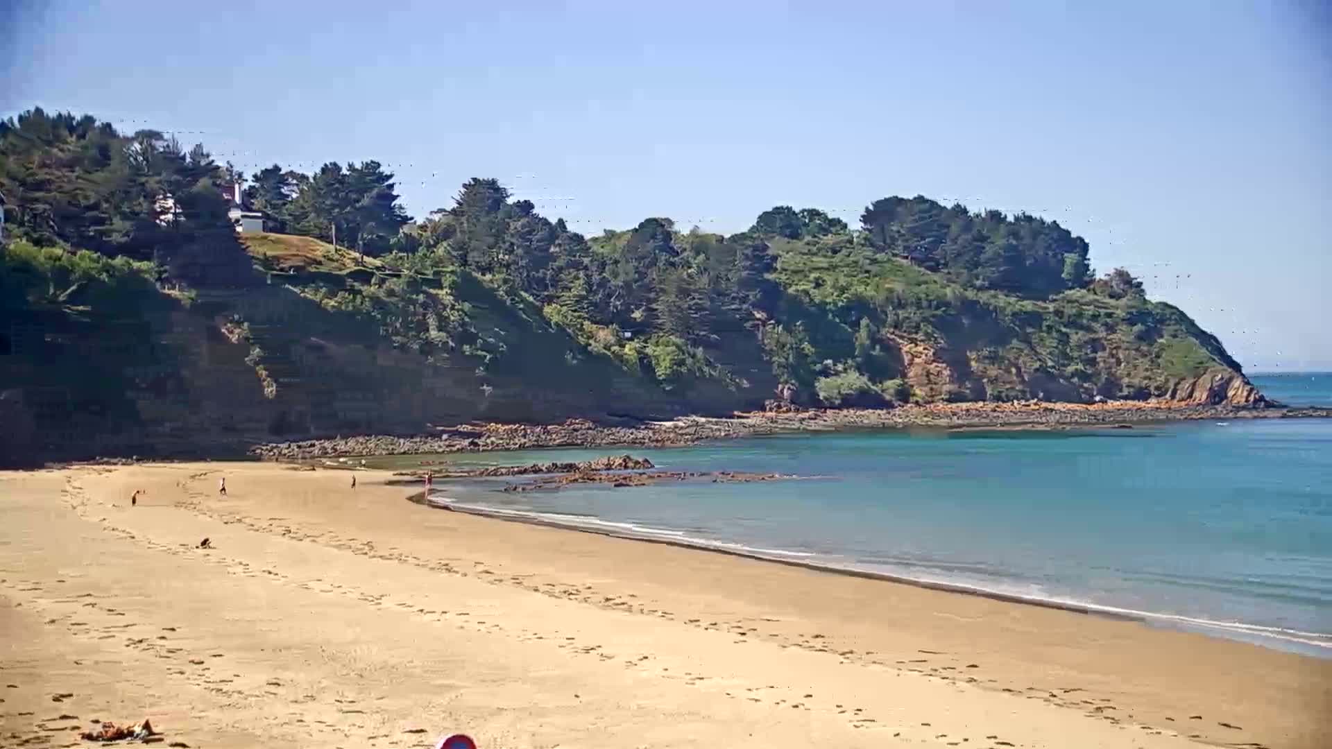 A sandy beach with a few people on it stretches to a calm, turquoise bay bordered by a verdant, tree-covered cliff on a sunny day.
