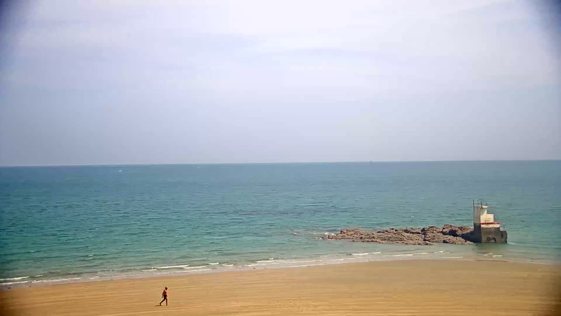 A person walks on a sandy beach next to a calm, teal ocean with a small structure on rocks just offshore under a mostly clear sky.