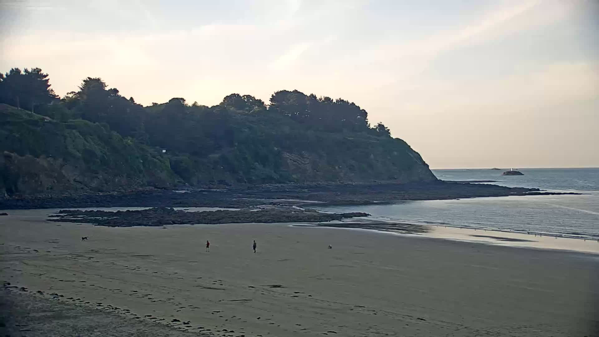 A sandy beach stretches out to a calm sea, bordered by a rocky shoreline and a verdant cliff topped with trees under a pale sky.