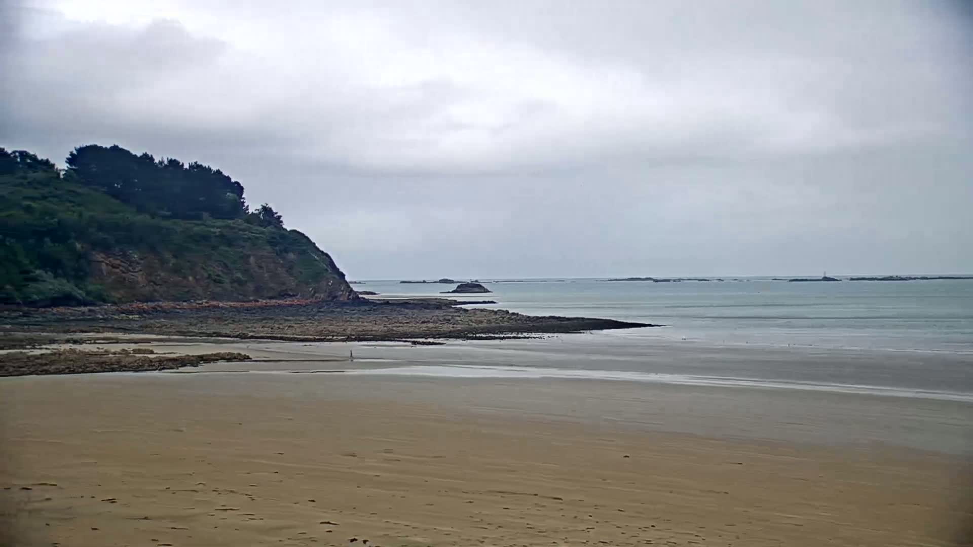 A sandy beach stretches out to a calm sea under an overcast sky, with a rocky shoreline and a green hill in the background.