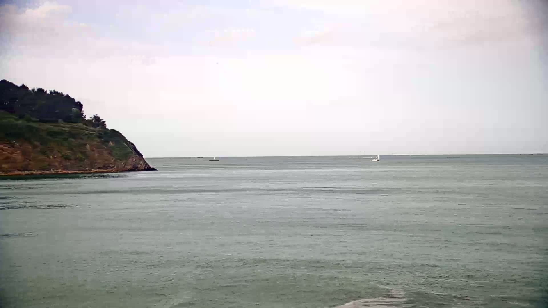 A calm, overcast day shows a green, tree-covered headland jutting into a body of water where a few sailboats are visible in the distance.