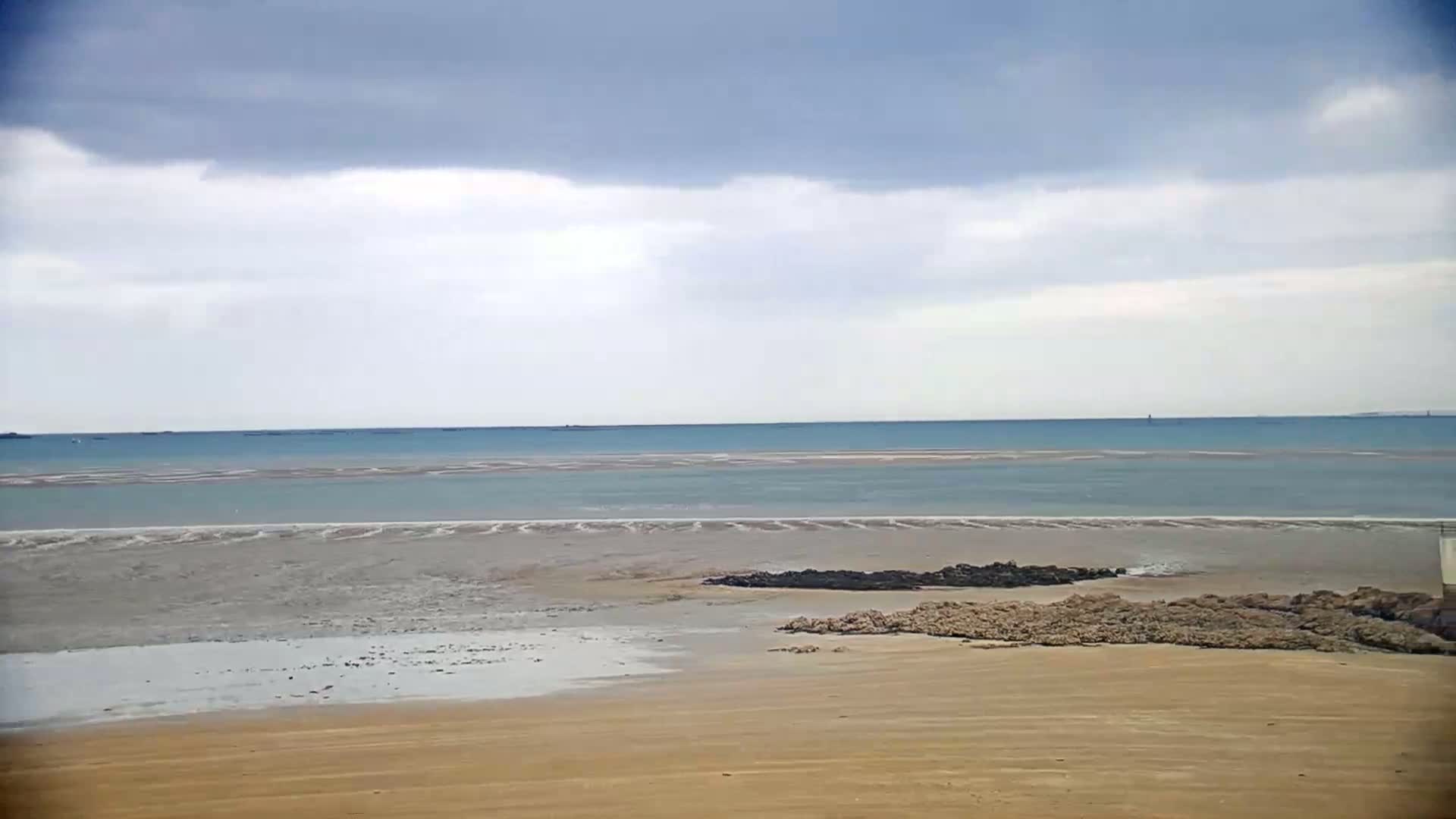 A sandy beach stretches to a calm, pale-blue sea under a mostly cloudy sky.