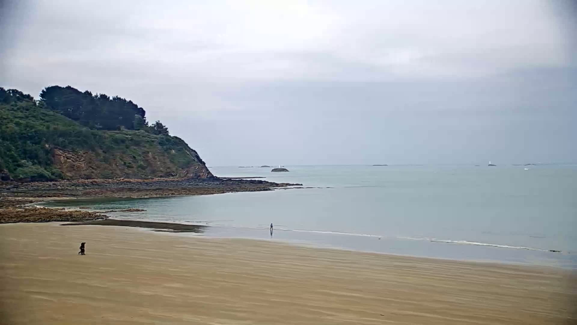 A sandy beach stretches out to a calm, light-grey sea under an overcast sky, with a small, green, rocky hill in the background and two people walking on the shore.