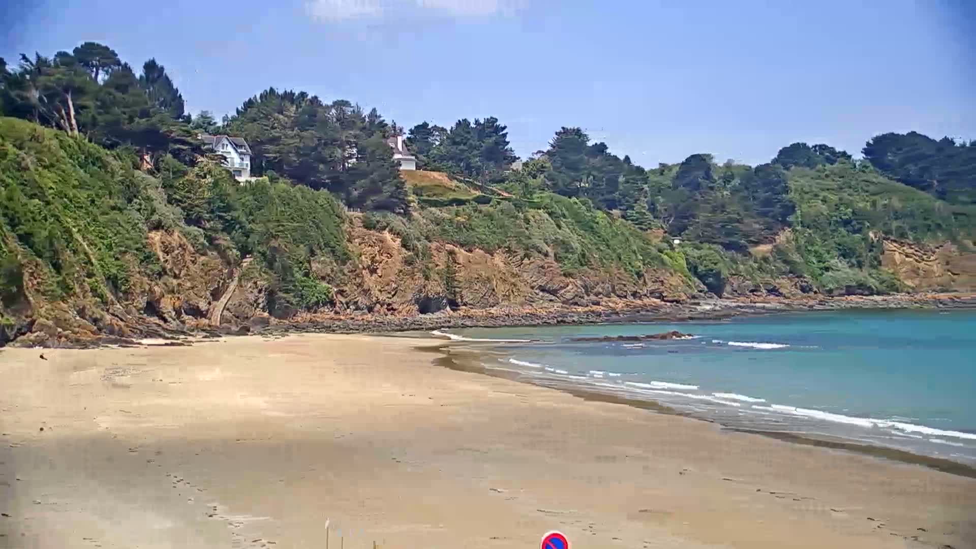 A sandy beach curves gently into a calm, turquoise bay, bordered by a verdant cliff topped with lush green trees under a clear blue sky.
