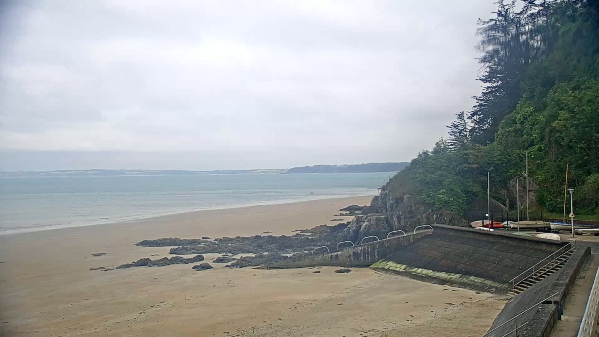 A sandy beach with dark rocks and a calm sea stretches to a distant shoreline under an overcast sky, next to a small boat ramp and lush green cliffs.