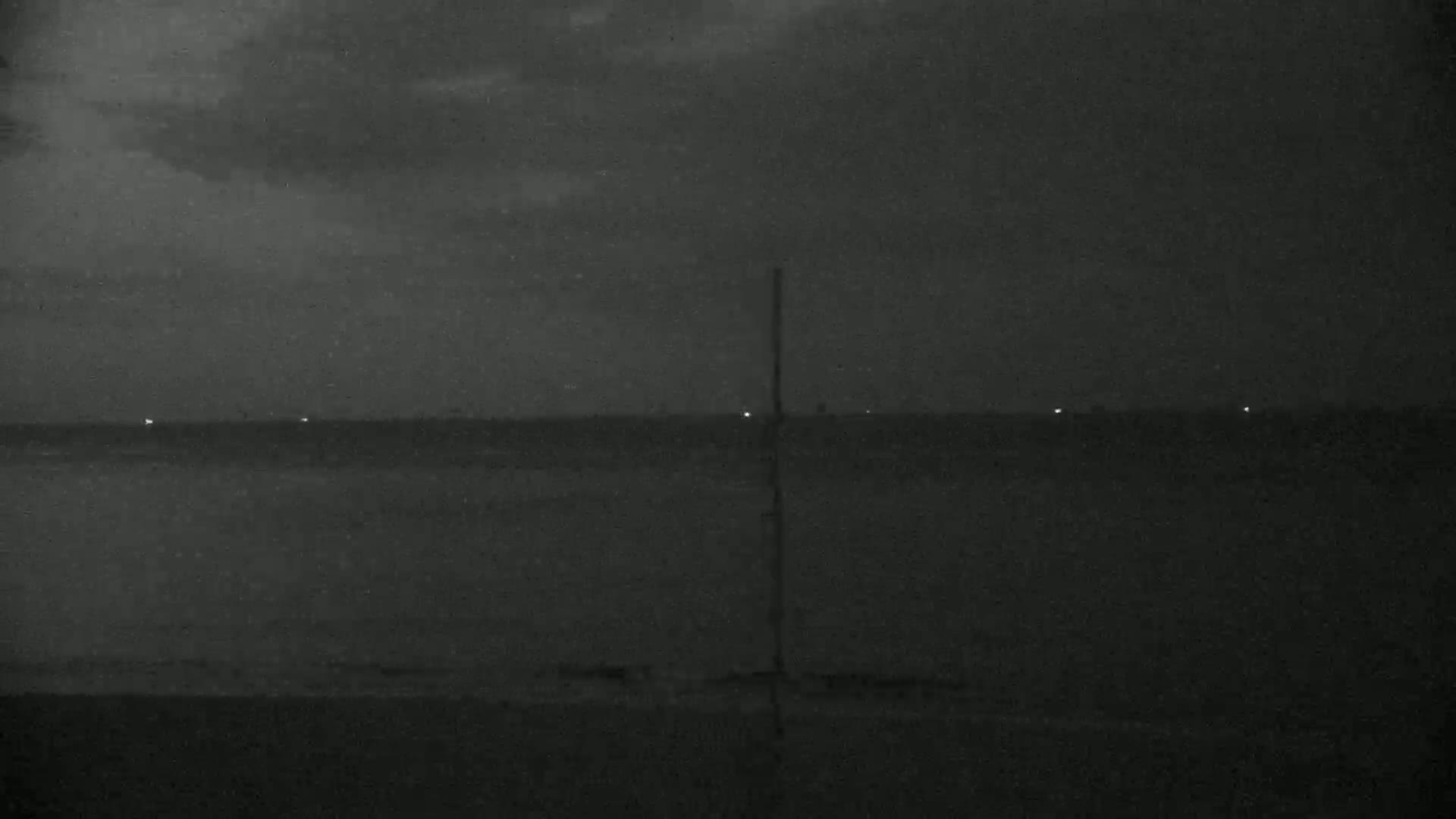 A dark, nighttime view of a calm body of water with a few faint lights on the horizon under a mostly cloudy sky.