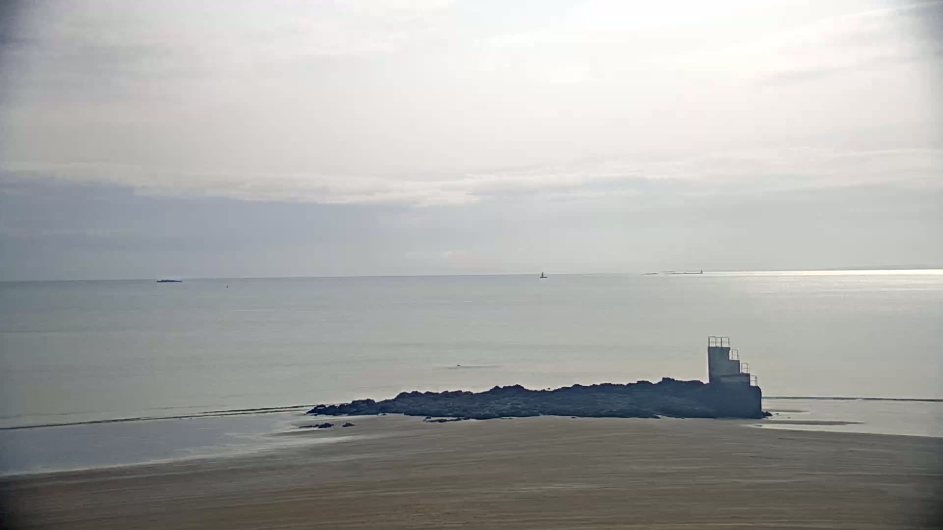 A sandy beach stretches to a calm sea under a mostly cloudy sky, with a small rocky outcrop and a diving platform visible near the shore.