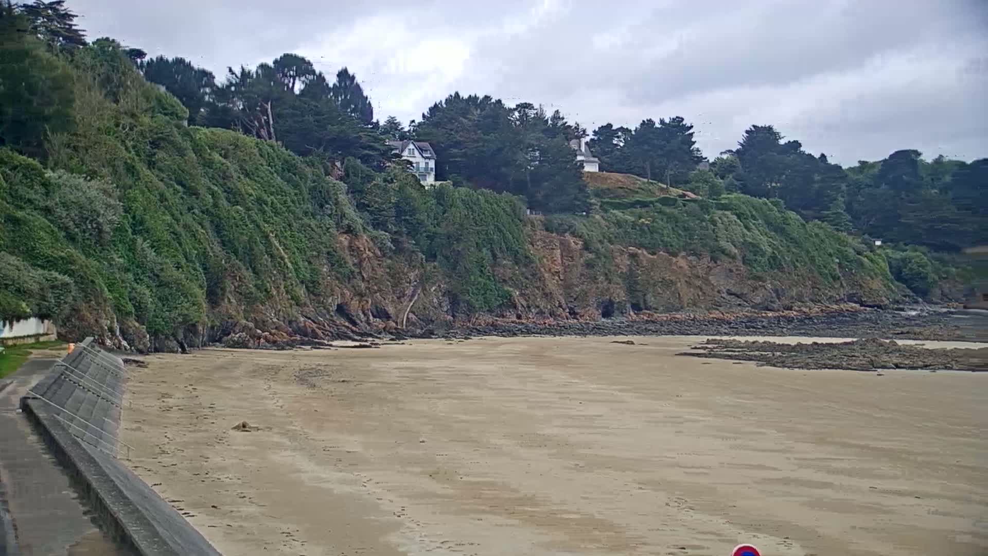 A sandy beach stretches out from a rocky, vegetated cliff under a cloudy sky.