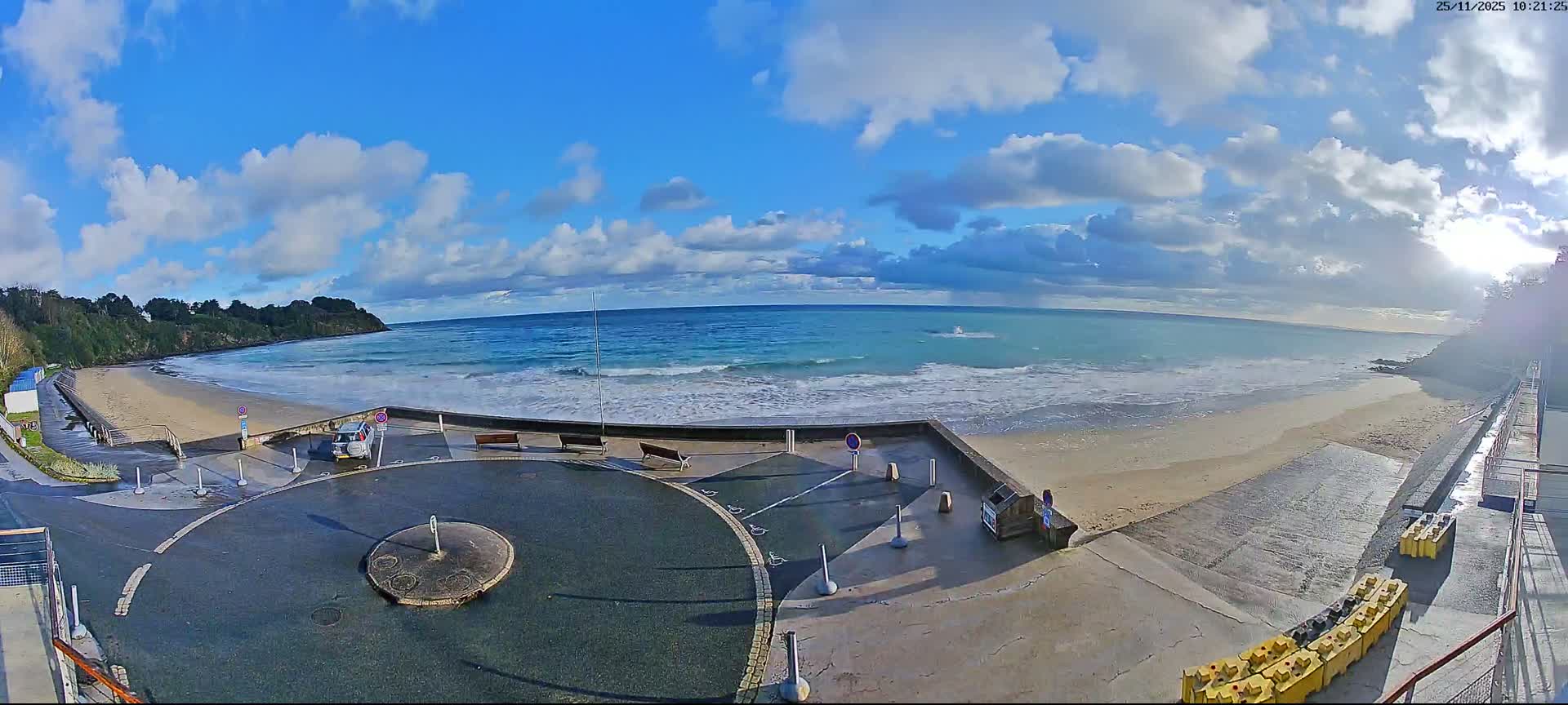The image displays a sunny, partly cloudy day over a wide sandy beach with active waves, flanked by green cliffs and a paved promenade featuring benches and a parked car.