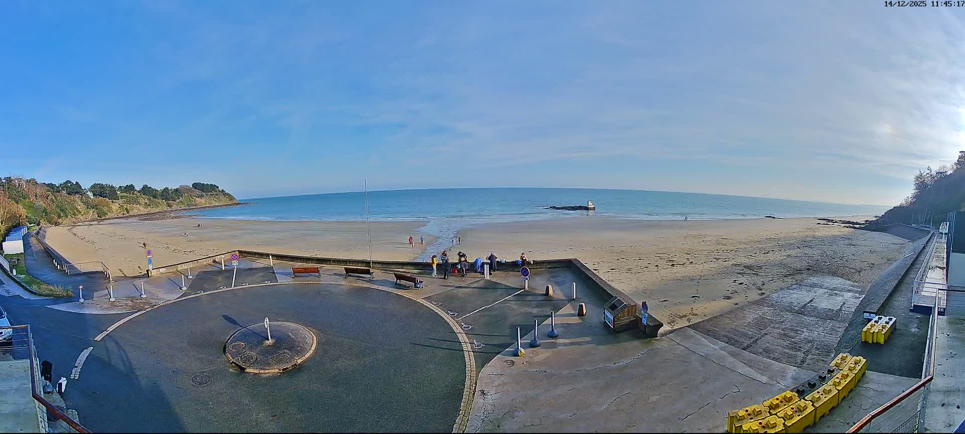 The image displays a sunny, partly cloudy day over a wide sandy beach with active waves, flanked by green cliffs and a paved promenade featuring benches and a parked car.