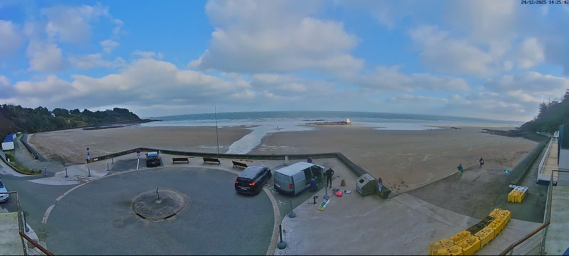 The image displays a sunny, partly cloudy day over a wide sandy beach with active waves, flanked by green cliffs and a paved promenade featuring benches and a parked car.