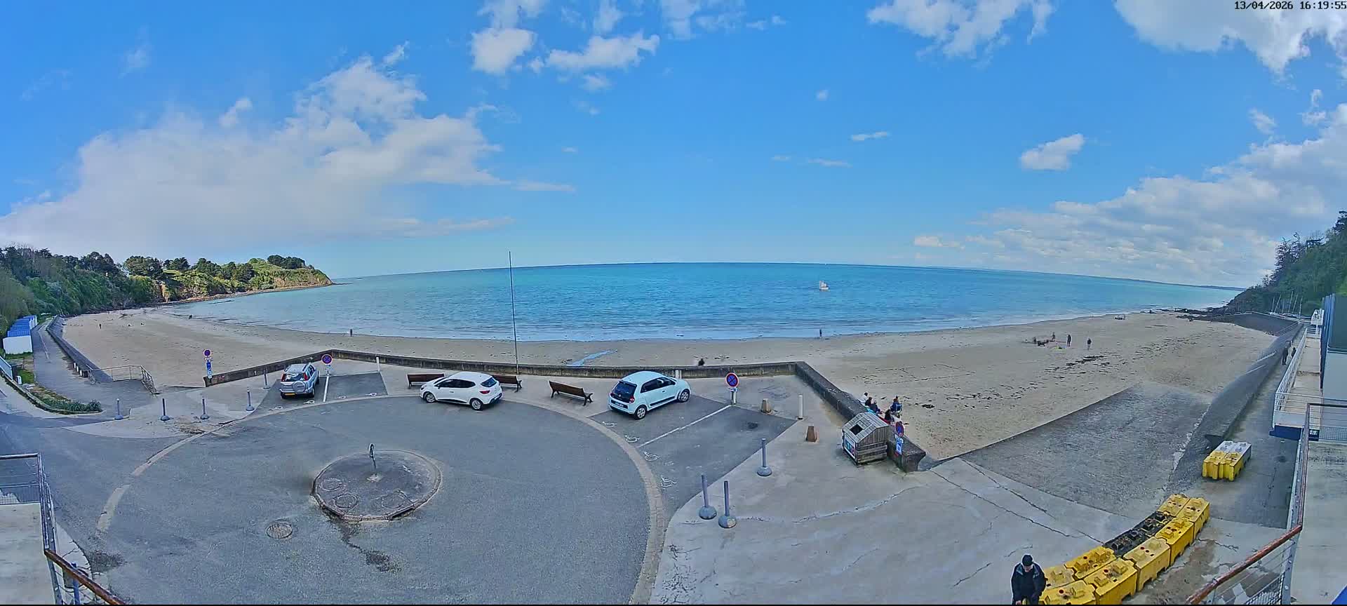 The image displays a sunny, partly cloudy day over a wide sandy beach with active waves, flanked by green cliffs and a paved promenade featuring benches and a parked car.