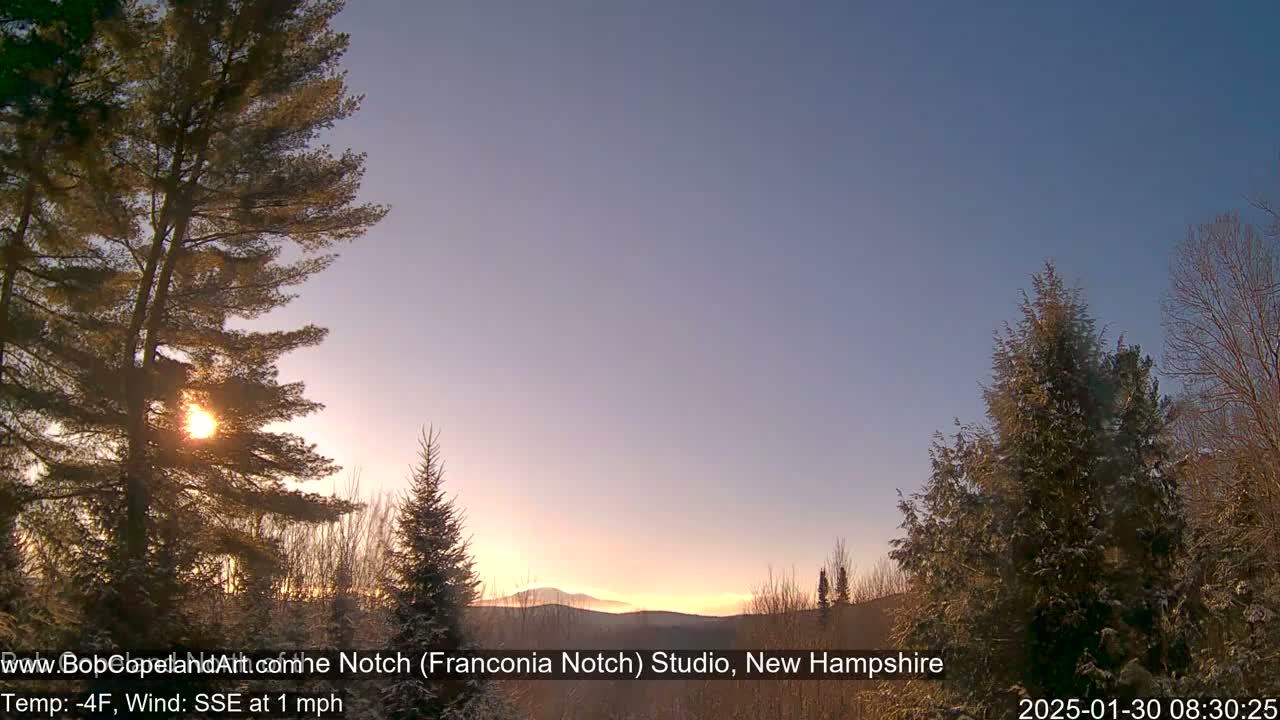 A sunrise casts a warm glow on a snowy landscape of evergreens and distant mountains, under a clear, cold sky.