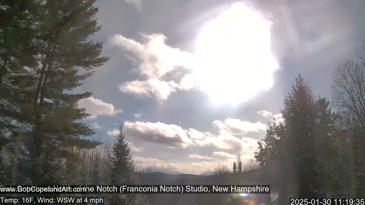 A bright, partly cloudy sky with the sun shining over a snow-covered landscape of evergreens and distant mountains.