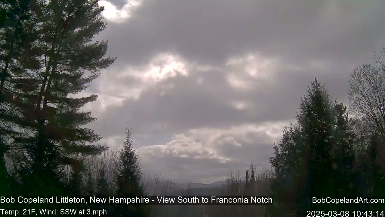 A mostly cloudy sky with a slight breeze, partially obscuring the sun, is visible over a winter landscape of evergreens.