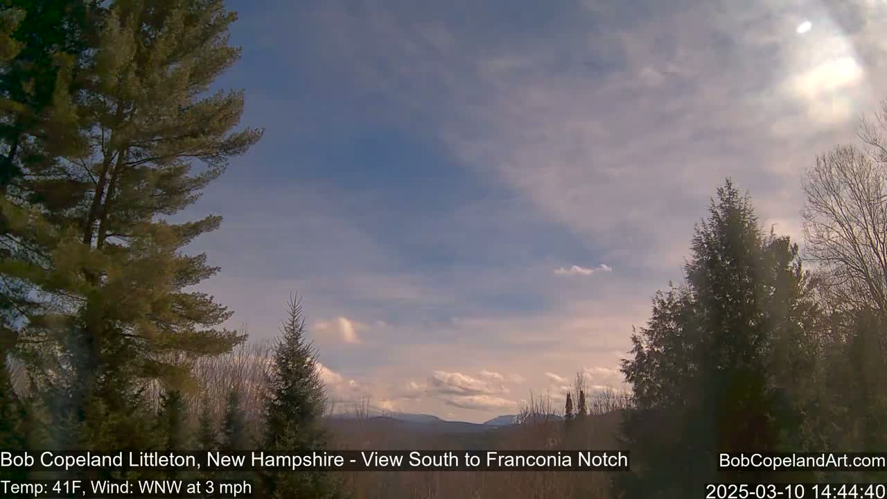 A partly cloudy sky over a winter landscape of evergreens and distant mountains.