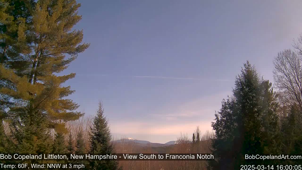 A clear, pale blue sky stretches over a landscape of evergreens and distant mountains under a light, nearly cloudless sky.