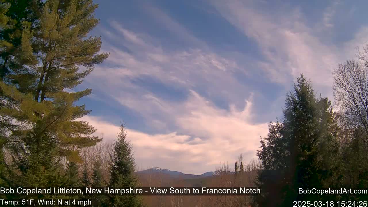 A partly cloudy sky with wispy clouds stretches over a mountainous landscape partially obscured by evergreens; a light north wind is blowing.