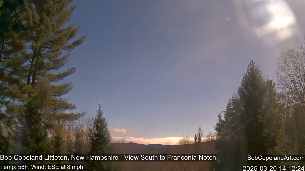 A mostly clear, sunny sky over a hilly landscape with evergreen and deciduous trees, under partly cloudy conditions with a light breeze.