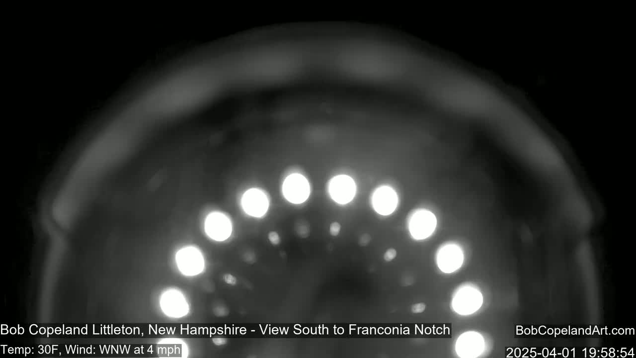 A nighttime, blurry, grayscale image shows a curved row of out-of-focus lights against a dark sky, with a temperature of 30°F and a wind of 4 mph from the west-northwest.