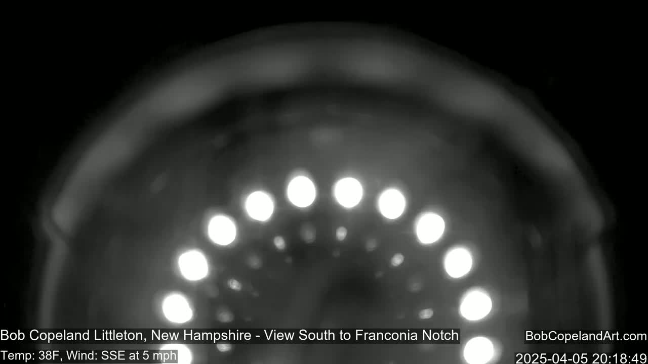 A nighttime, blurry, grayscale image shows a semi-circular arrangement of bright lights against a dark background, with a 38°F temperature and a 5 mph southeasterly wind.