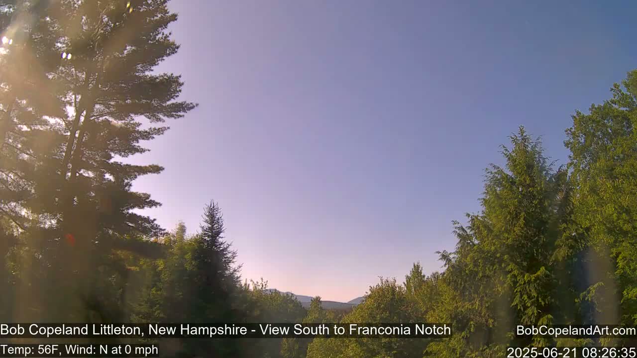 A mostly clear, calm sky with a slight haze is visible over a green, wooded landscape, sunlit from the left.
