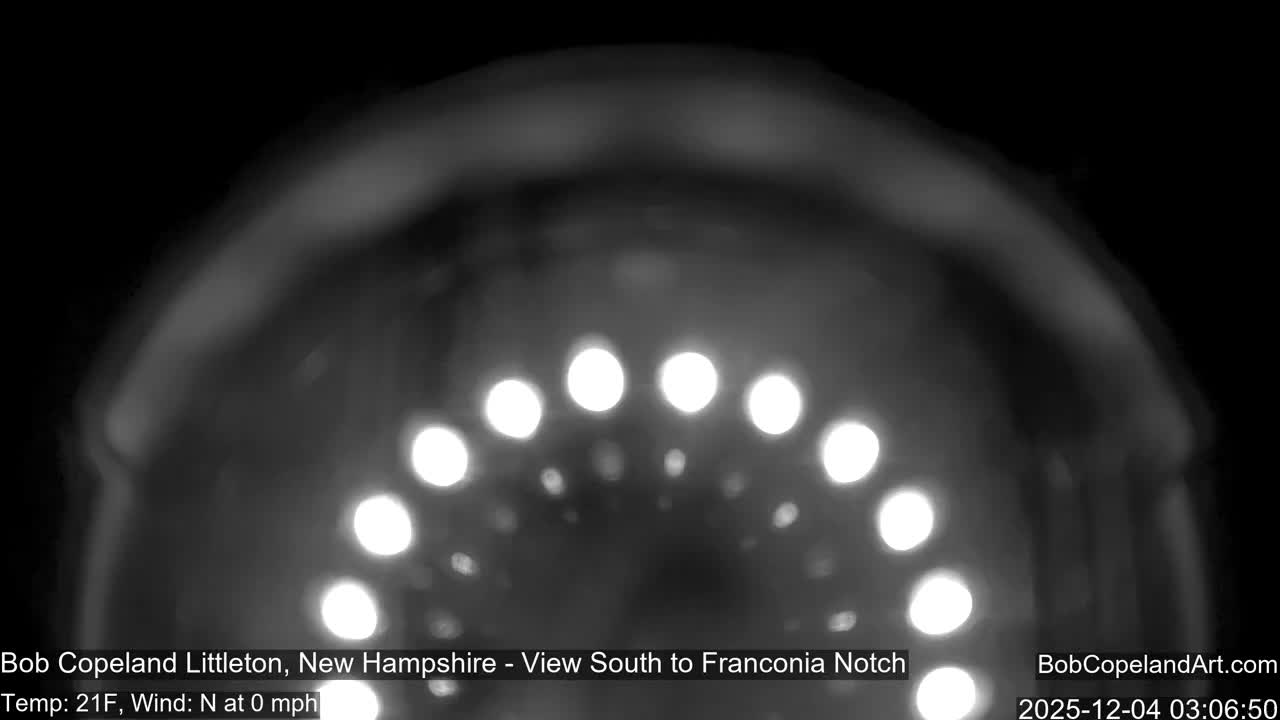 A blurry, monochromatic nighttime outdoor scene displays an arc of bright, circular lights through what appears to be a camera dome, with a temperature of 21F and no wind.