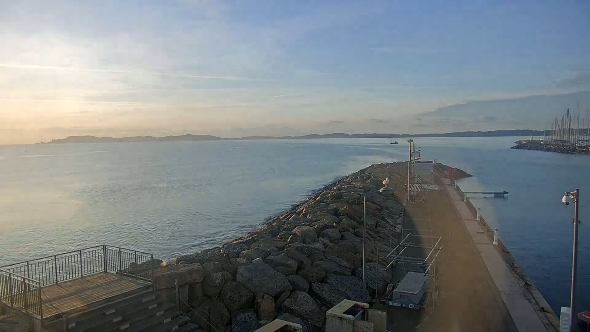 A calm, sunrise-lit body of water stretches to a distant landmass, seen from a rocky breakwater with a paved walkway leading to a small harbor.