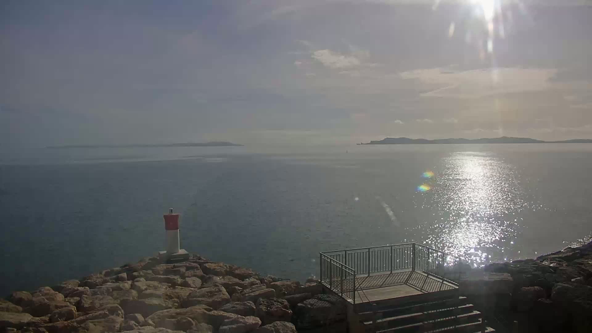 A rocky shoreline with a small viewing platform overlooks a calm, sunlit sea, distant islands visible on the horizon under a partly cloudy sky.