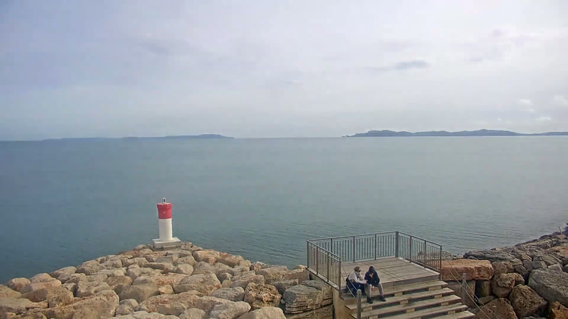 Two people sit on a wooden deck overlooking a calm sea with a small, red and white lighthouse on a rocky outcrop under a lightly cloudy sky.