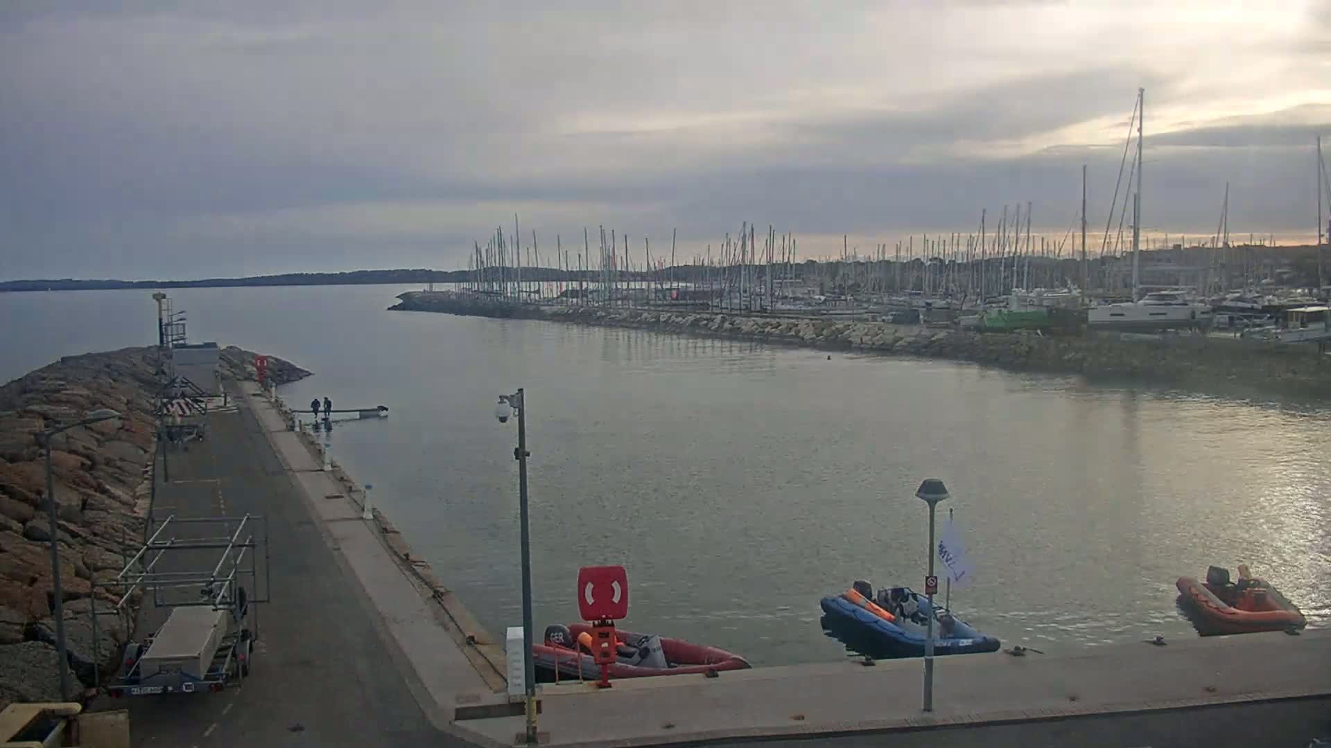 A calm harbor filled with numerous sailboats under an overcast sky, with several small motorboats docked near a rocky pier.