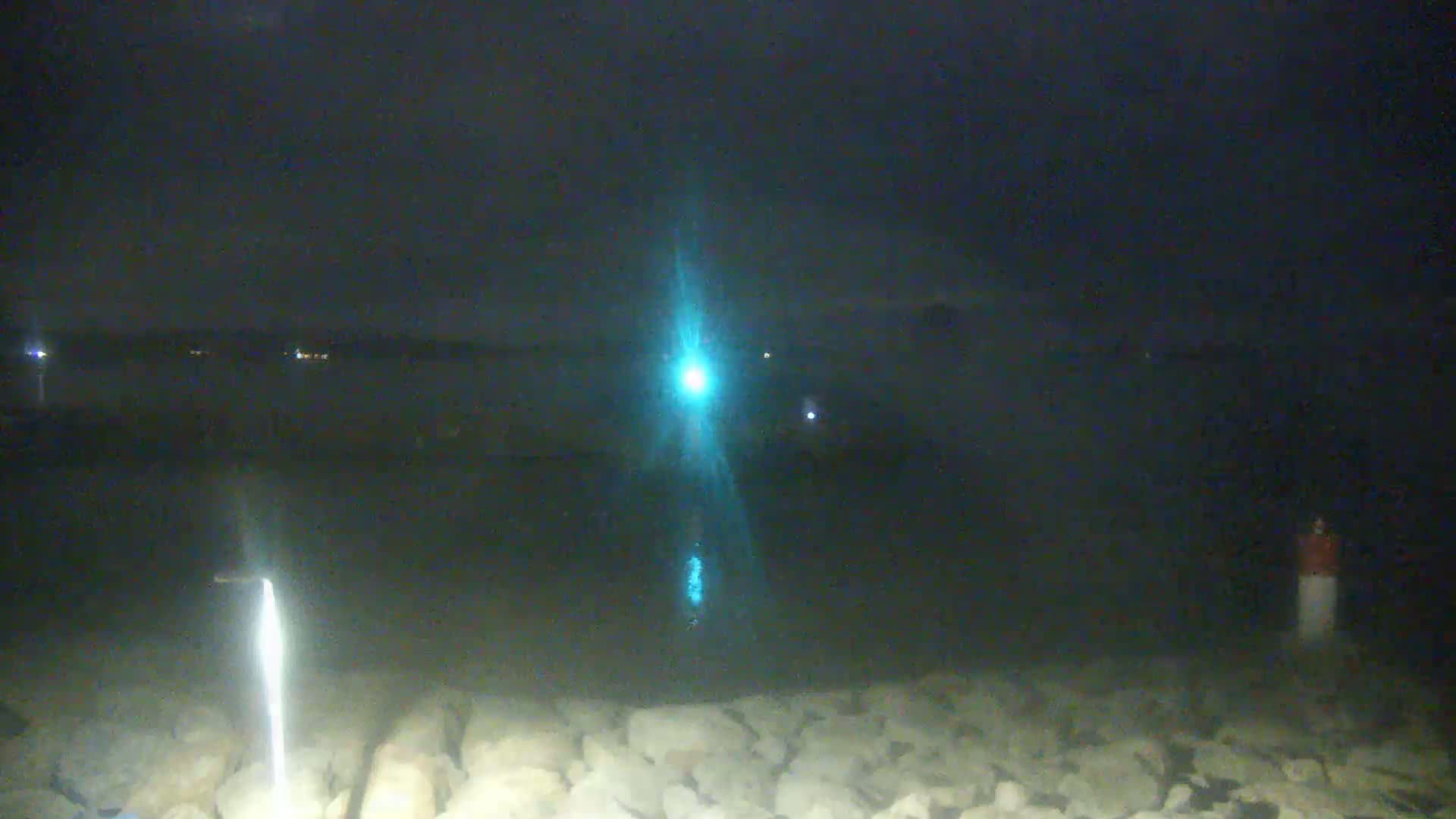 A nighttime view of a rocky shoreline with a bright light reflecting off the dark water.