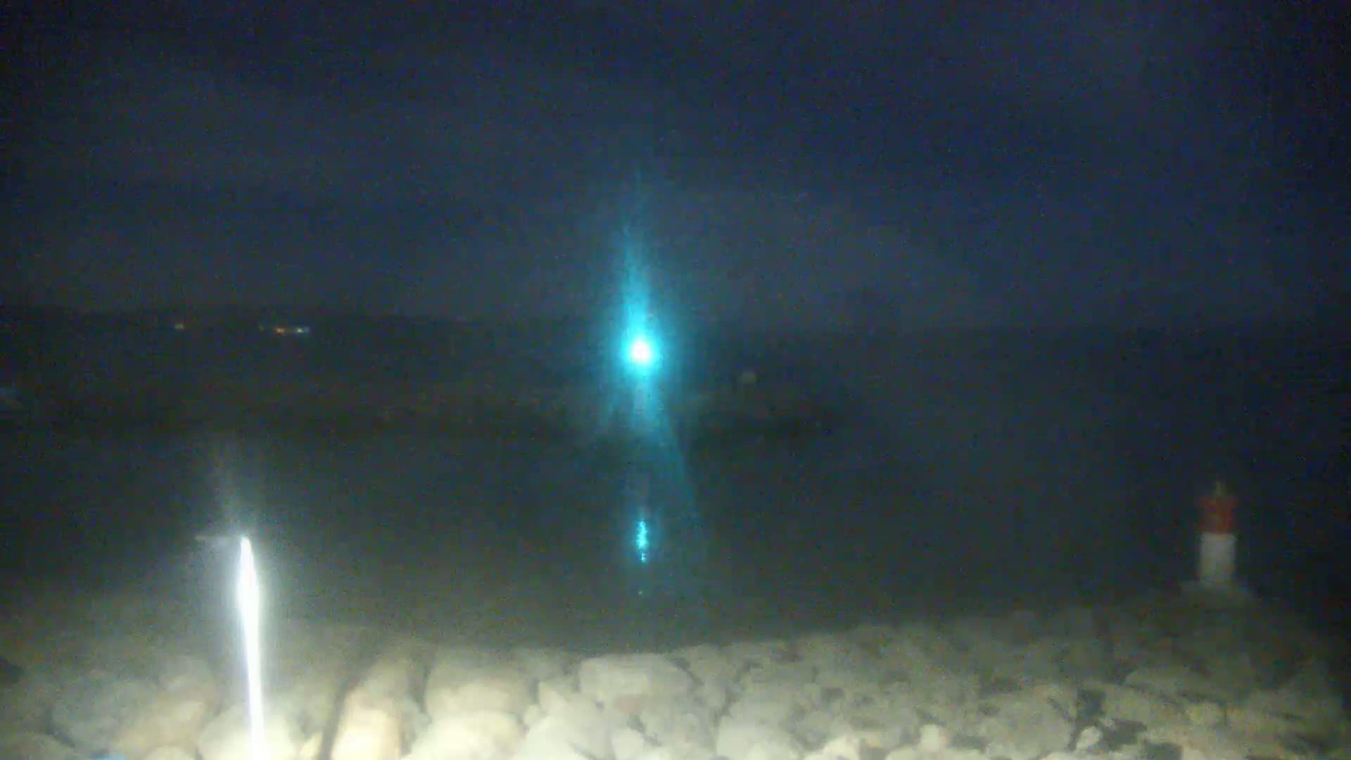 A bright teal light shines over dark water from a distance, illuminating a rocky shoreline at night.