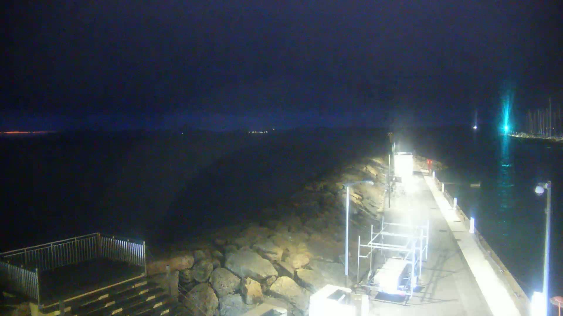 A dark, nighttime view of a paved walkway along a rocky waterfront, under a mostly clear sky.