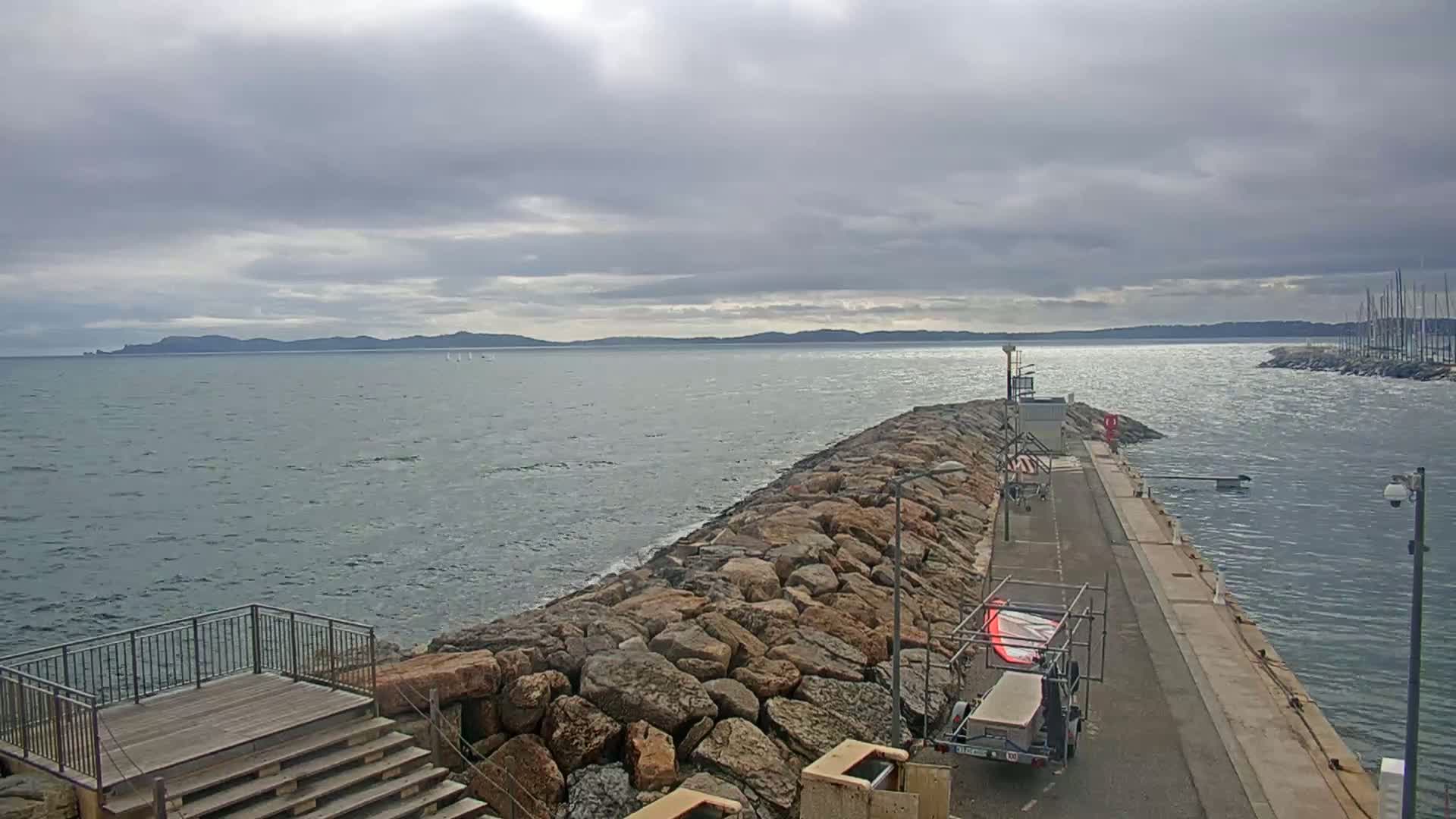 A rocky breakwater extends into a choppy sea under a cloudy sky, with a distant landmass visible and a few sailboats in the water.